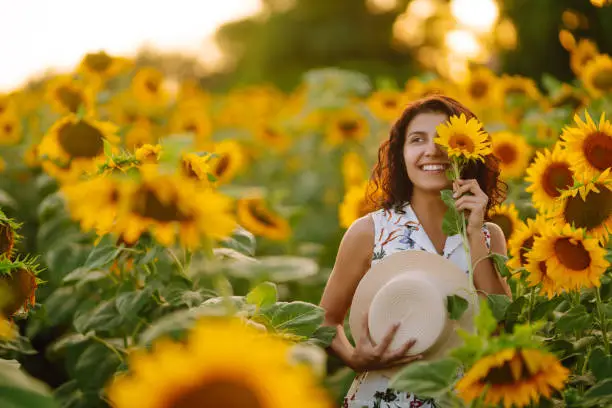 Young woman walks  on blooming sunflower field. Happiness with nature. Beautiful woman posing in a field of sunflowers in a dress and hat at sunset. Summer holidays, vacation, relax and lifestyle.