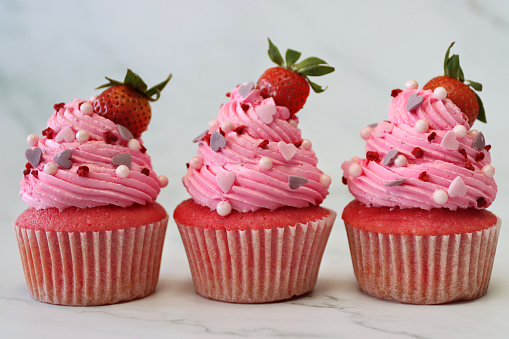 Photo showing a marble cake stand containing freshly baked, homemade, pink velvet strawberry cupcakes in paper cake cases. The cup cakes have been decorated with a swirl of pink piped icing, sugar craft hearts and sphere sprinkles topped with strawberries.