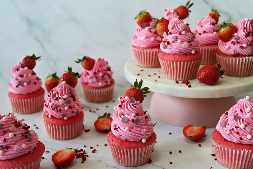 Photo showing a cake stand of freshly baked, homemade, pink velvet strawberry cupcakes in paper cake cases. The cup cakes have been decorated with swirls of pink piped icing, sugar craft hearts and sphere sprinkles topped with a strawberry.