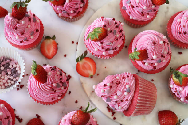 imagem de lote de cupcakes caseiros de veludo rosa de morango em caixas de bolo de papel, morango em redemoinhos de cobertura de manteiga rosa, açúcar artesanal forma coração e polvilho esfera, um cupcake deitado ao lado, visão elevada - cupcake sprinkles baking baked - fotografias e filmes do acervo