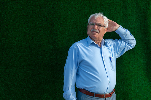 A portrait of 60-69 years old Turkish mature man. He put his elbow against the green colored wall. He puts his hand on his face and thinks some things. He has a mustache and looks away.