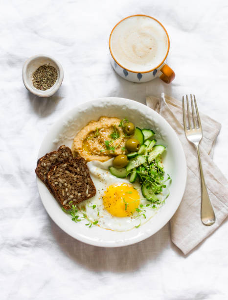 istock Delicious breakfast, brunch - fried egg, fresh vegetables, hummus, olives, micro greens, grain bread and cappuccino on a light background, top view 1320623035