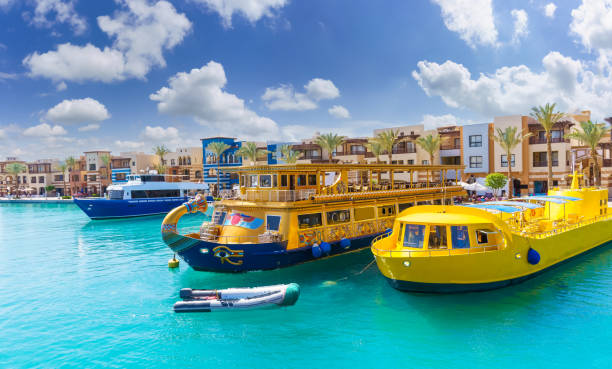A white boat resting in a harbor, surrounded by other boats in the background. The boat appears to have suffered damage and is partially submerged in the water. It has a flat roof and a cabin area that has collapsed partially. The boat leans to the left, and its front is submerged in the light blue water. The sky above is a hazy shade of blue, adding to the overall scene.