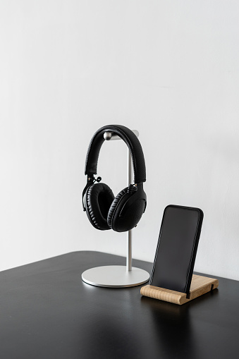 Vertical shot of black wireless headphones on metal stand next to smartphone with blank display at dark table surface against white copy space wall. Modern technologies concept