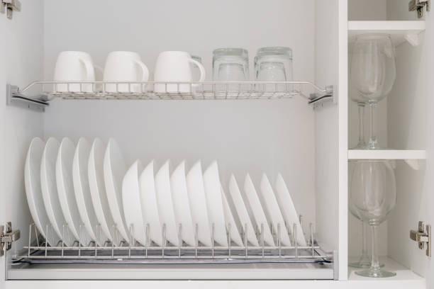 Set of clean white dishes, mugs and different glasses and other crockery in opened white kitchen cupboard shelf. Close up of storage cabinet with kitchenware inside
