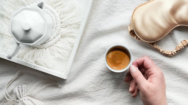 istock Espresso in hand, tray with white ceramic espresso coffee maker on trendy macrame pad. Sleep mask on crumpled bed sheets. Female hand with cup of espresso. Good morning Simple minimal top view 1320651695