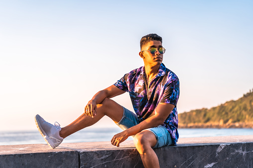 Summer lifestyle, portrait of a young Latino man enjoying summer vacation. with unbuttoned purple flower shirt and sunglasses