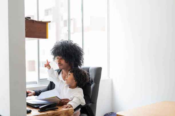 istock Young black father with his Latino daughter both sitting at the workplace while the father looking to finish his work and his daughter distracts him by playing 1320657604