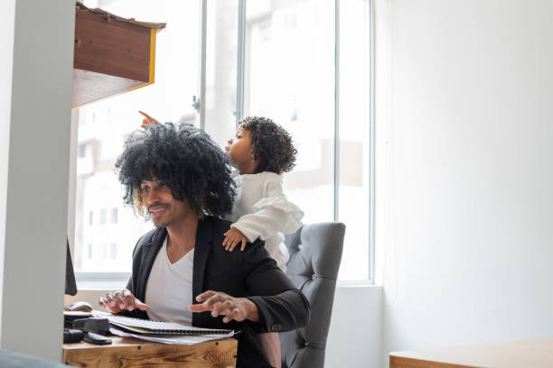 istock Young black father with his Latino daughter both sitting at the workplace while the father looking to finish his work and his daughter distracts him by playing 1320658221