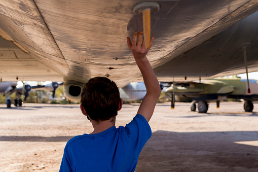 A boy is walking underneath the fuselage of a large airplane, holding up his hand to touch the surface of the plane.  He admires the size and engineering of the machine.