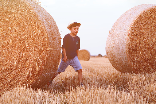 Young cute country boy in hat near haystacks at sunset in summer. Summer concept, a vacation. Active outdoors leisure with children on warm summer day.