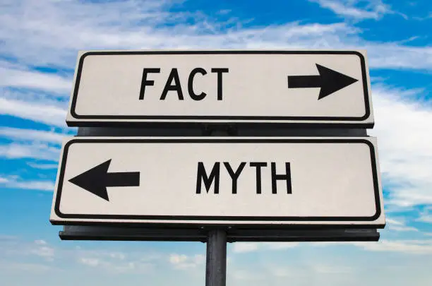 Photo of Fact versus myth road sign with two arrows on blue sky background. White two street sign with arrows on metal pole. Two way road sign with text.