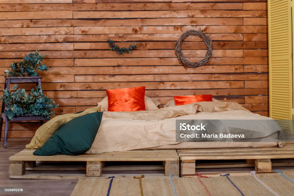Pallet bed covered with a beige blanket with two red pillows Pallet bed covered with a beige blanket with two red pillows, against the background of a wall of wooden planks Bed - Furniture Stock Photo