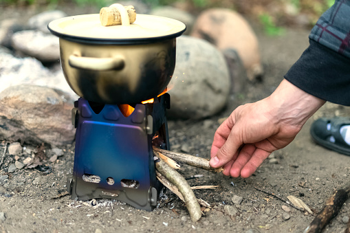 A small beige saucepan with a lid is simmering on a fire, cooking in a camping