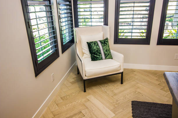 Modern automatic roller blinds and sun shades cover large windows, diffusing natural light into cozy room. Blackout and screen fabrics close-up. Space features stylish chair and minimalist side table.
