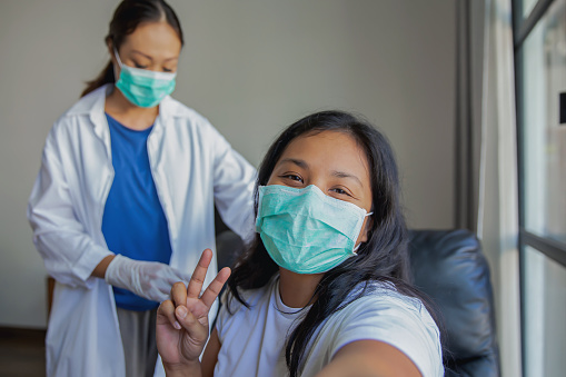 Close up shot of happy Malaysian woman with face mask , taking a selfie before vaccination. She's smiling behind her mask and posing a peace sign gesture to the camera.Behind her there's a female doctor preparing the injection of the Covid-19 vaccine.