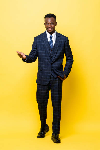 portrait en pied d’un jeune bel homme africain en costume d’affaires souriant et ouvrant sa paume dans un fond de studio isolé jaune - lenght photos et images de collection
