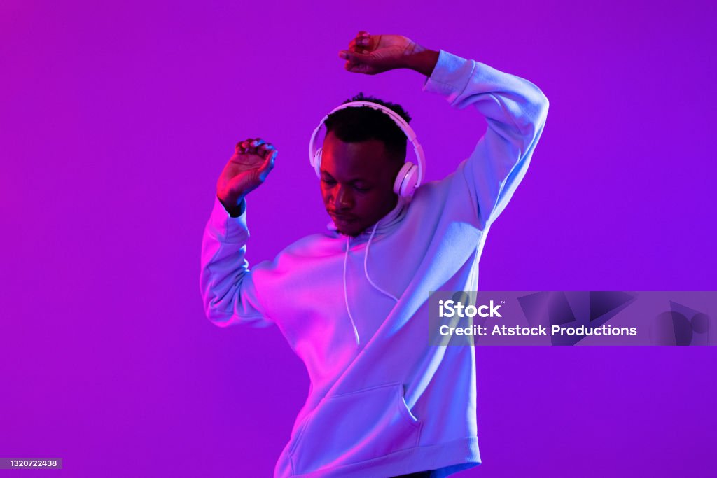Young handsome African man wearing headphones listening to music and dancing in futuristic purple cyberpunk neon light background Music Stock Photo