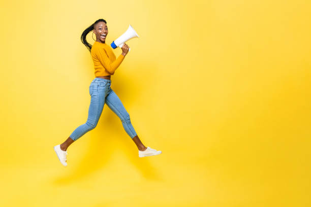 retrato saltartivo de una joven afroamericana sonriente extática con megáfono en un fondo amarillo de estudio aislado con espacio de copia - contar fotografías e imágenes de stock