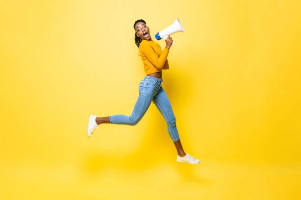 孤立したスタジオ黄色の背景で発表メガホンを保持しているショックを受けた若いアフリカ系アメリカ人女性のジャンプ肖像画 - 語る ストックフォトと画像