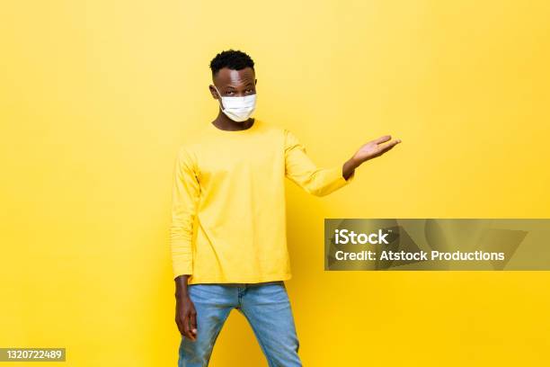 Portrait Of Young African Man Wearing Face Mask Protecting From Corovavirus With Hand Opening To Empty Space Aside In Isolated Yellow Studio Background Stock Photo - Download Image Now