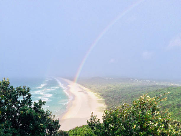 Horizontal landscape photo of a rainbow arching over the coastal forest, sand dunes and beach at popular surfing spot, Tallow Beach, Byron Bay
