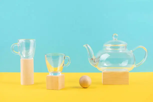Empty glass transparent tea set. Teapot and and two cups standing on wooden geometric shapes, yellow and blue background. Front view.