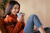 セーターを着たアジアの若い女性の肖像画。彼女は香りを吸い込み、冬の朝のコーヒーを飲んだ。彼女は笑顔で、家でリラックスして楽しんでいます。