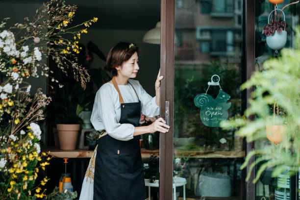 若いアジアの女性の花屋、彼女のビジネスの日を開始する中小企業の花屋の所有者。ドアを開けて、彼女の店で開いた看板をぶら下げる。中小企業のコンセプト。日常業務 - store opening ストックフォトと画像