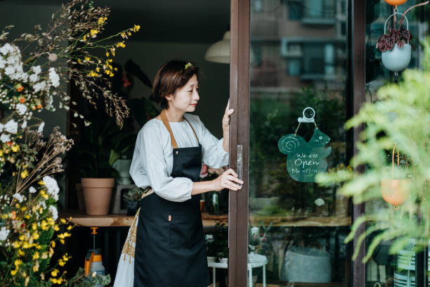 若いアジアの女性の花屋、彼女のビジネスの日を開始する中小企業の花屋の所有者。ドアを開けて、彼女の店で開いた看板をぶら下げる。中小企業のコンセプト。日常業務 - florist small business flower shop owner ストックフォトと画像