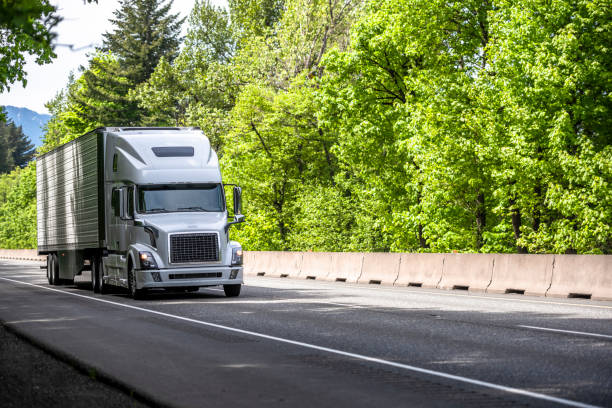 istock Bonnet white big rig semi truck tractor with turned on headlight transporting loaded refrigerator semi trailer moving on the green forest road 1320746068