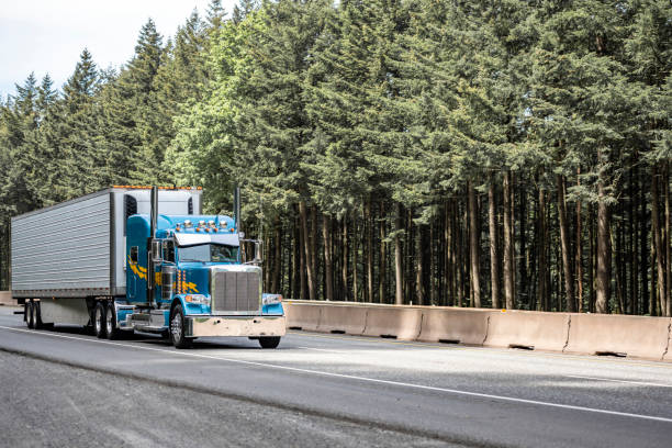 istock Classic blue big rig semi truck with custom chrome accessories transporting loaded refrigerated semi trailer with refrigerator unit on the front wall driving on the road with conifer trees on the side 1320748307