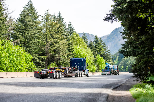 istock Team of two blue big rig semi trucks with step down semi trailers transporting transport equipment running at the convoy on the green forest road 1320750069