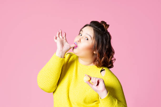 feliz modelo de talla grande comiendo macarrones, chica gorda ama dulces, fondo de estudio rosa - finger in mouth fotografías e imágenes de stock