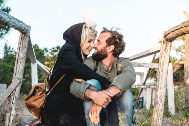 romantisches glückliches paar in der liebe sitzen auf holztreppe auf dünen am wilden strand bei sonnenuntergang - freund und freundin blickt in die augen des anderen - reisende verliebt - naturliebhaber konzept - backpacker green vacations outdoors stock-fotos und bilder