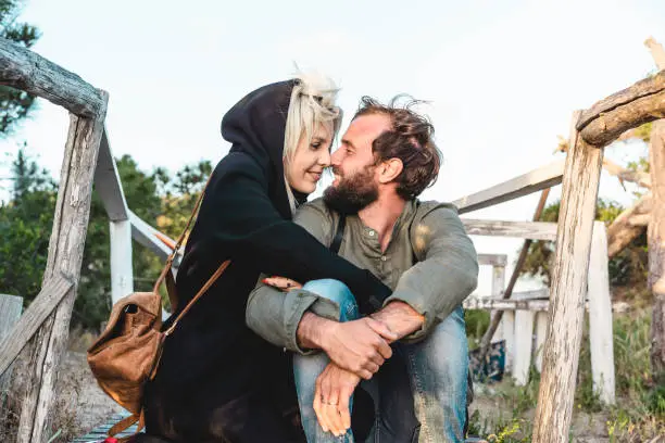 Photo of Romantic happy couple in love sitting on wooden stairs on dunes at the wild beach at sunset - Boyfriend and girlfriend looking into each other's eyes - Travellers in love - nature lovers concept