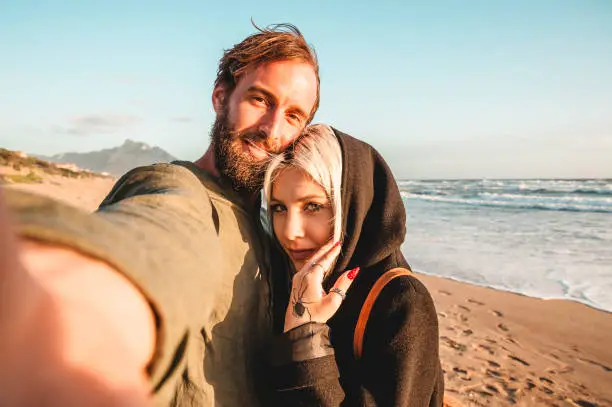 Traveling couple in love taking selfie in a deserted beach at sunset - Woman with magnetic gaze and her hipster boyfriend having fun while looking camera for selfie - Black spider tattoo on woman hand