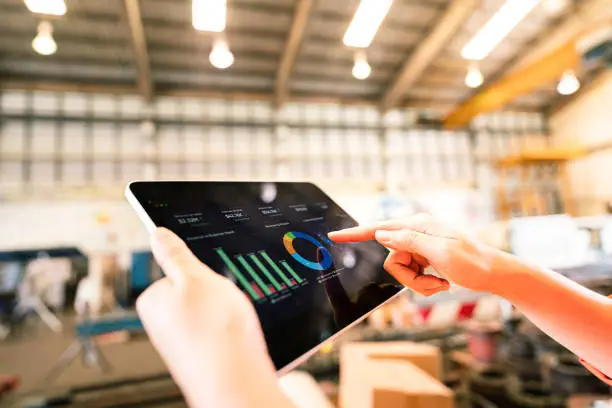 Action of a worker is using tablet to review storage report with blurred background of warehouse. Business management and technology concept photo. Close-up and selective focus at human's hand part.