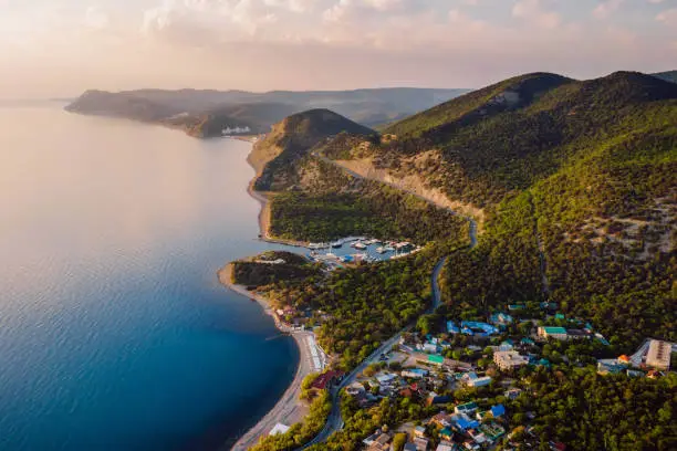 Aerial view of coastline with blue sea, mountains with trees, cliffs with sunset light.