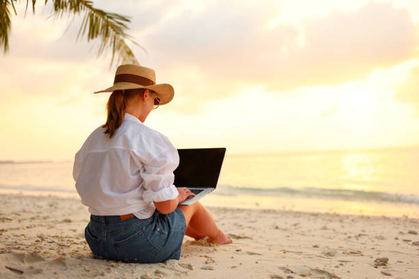 istock Young female freelancer wearing straw hat working on laptop while sitting on tropical beach at sunset 1320766094