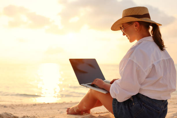 istock Young female freelancer wearing straw hat working on laptop while sitting on tropical beach at sunset 1320766214