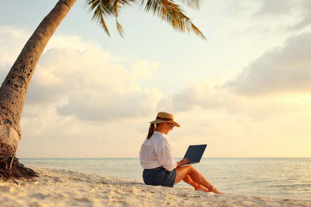 istock Young female freelancer wearing straw hat working on laptop while sitting on tropical beach at sunset 1320766290