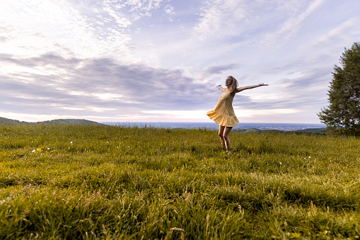 Young woman in white dress stands in the middle of green meadow. Enjoying nature and free life. Selective focus.