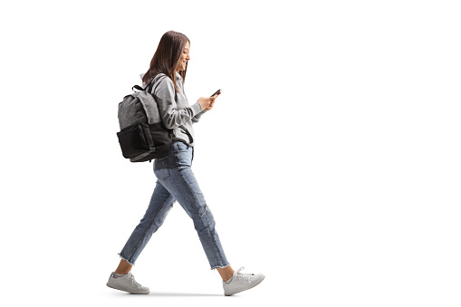 Full length profile shot of a female student walking with a mobile phone isolated on white background