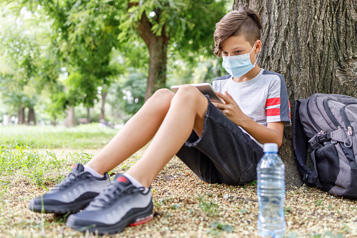 Boy with protective mask on his face  relaxing by a tree in park and using dgital tablet