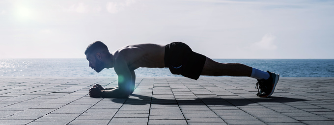 Fit young man doing plank on seafront. Fitness training in morning outdoors. Workout during lockdown outside the gym.