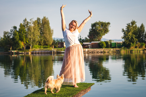 Happy woman have fun with her cairn terrier dog on pier at lake during hot summer day. Smiling plus size woman with arms raised