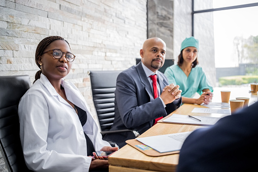 Young female doctor sitting in meeting at hospital conference room. Hospital staff in boardroom meeting listening to administrator.