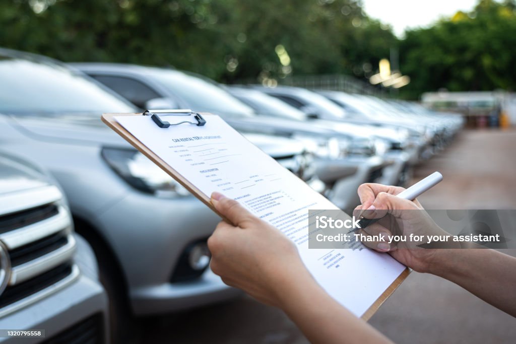 Signing on the agreement term of car rental service - Business and transportation service concept. Action of a customer is signing on the agreement term of car rental service. Close-up and selective focus a human's hand with blurred background of cars in row. Business and transportation concept. Fleet of Vehicles Stock Photo