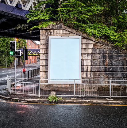 An empty billboard poster frame in vertical format next to a pavement which leads under an urban railway bridge.
